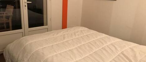 Quarto com colcha branca, porta de correr de vidro e um quadro na parede.