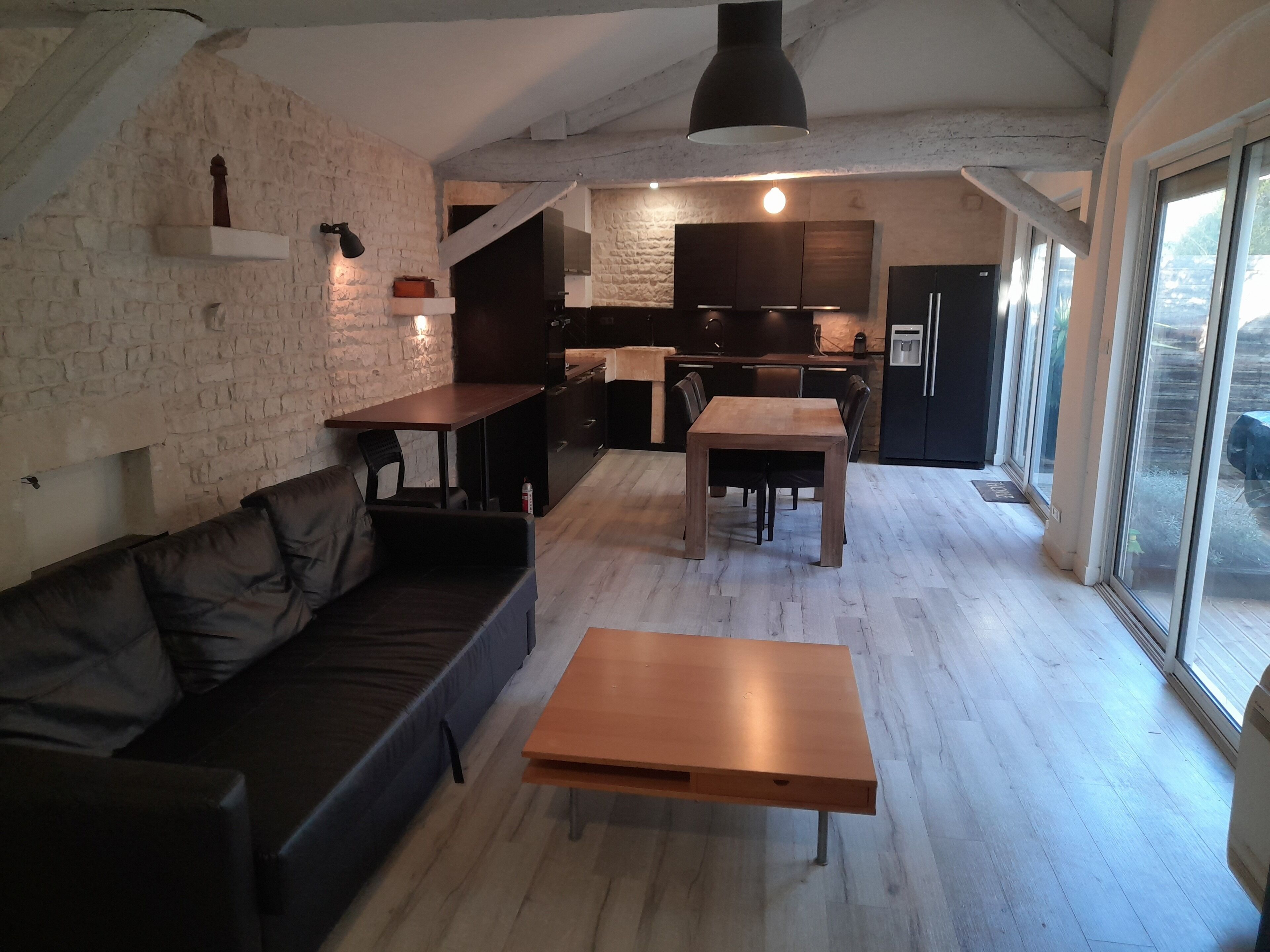 A spacious living area with a wooden table and sofa, a kitchen in the background, and large windows.