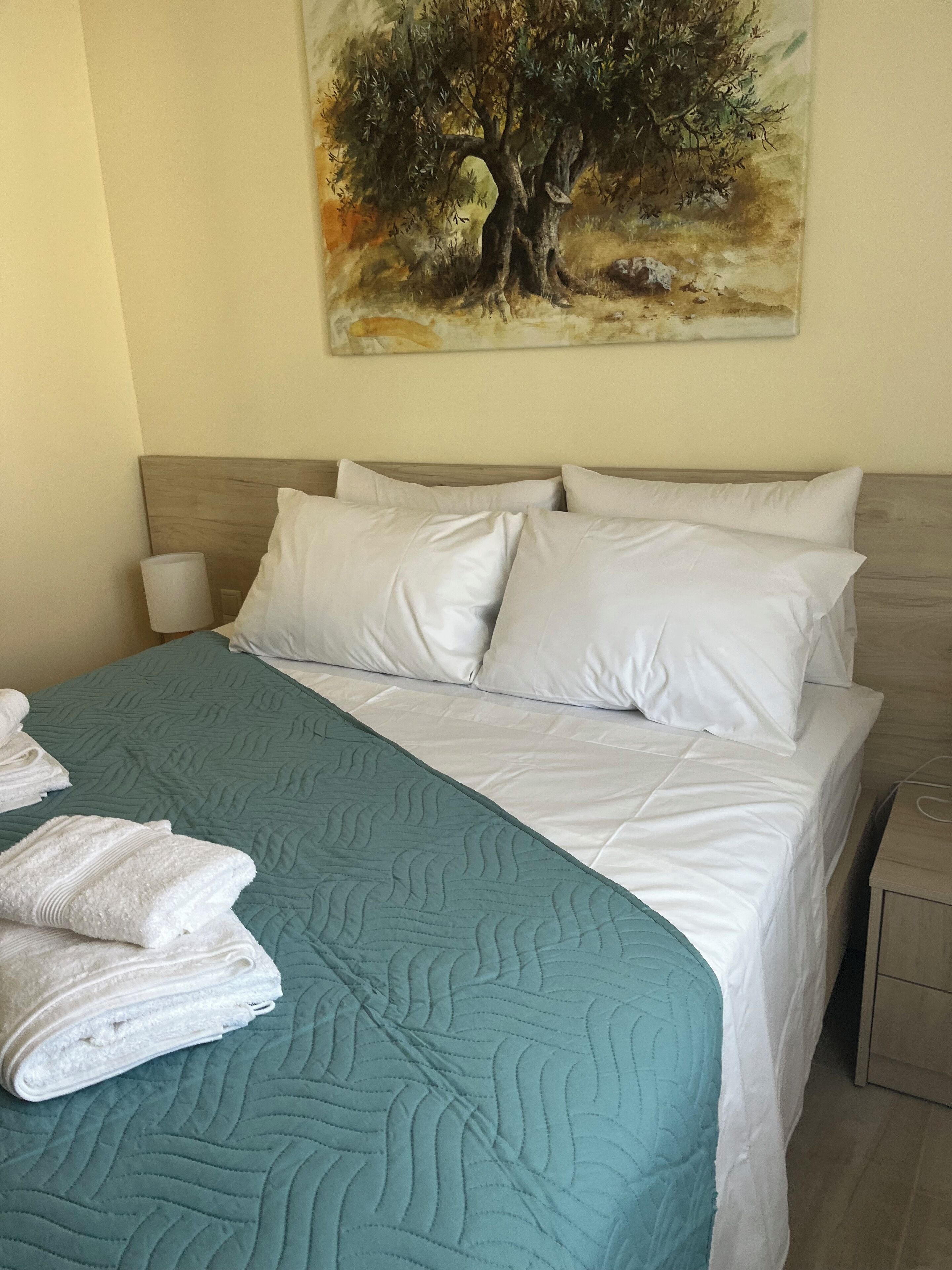 A neatly made bed with white linens and a teal quilt, a bedside lamp, and a folded towel on the bed.