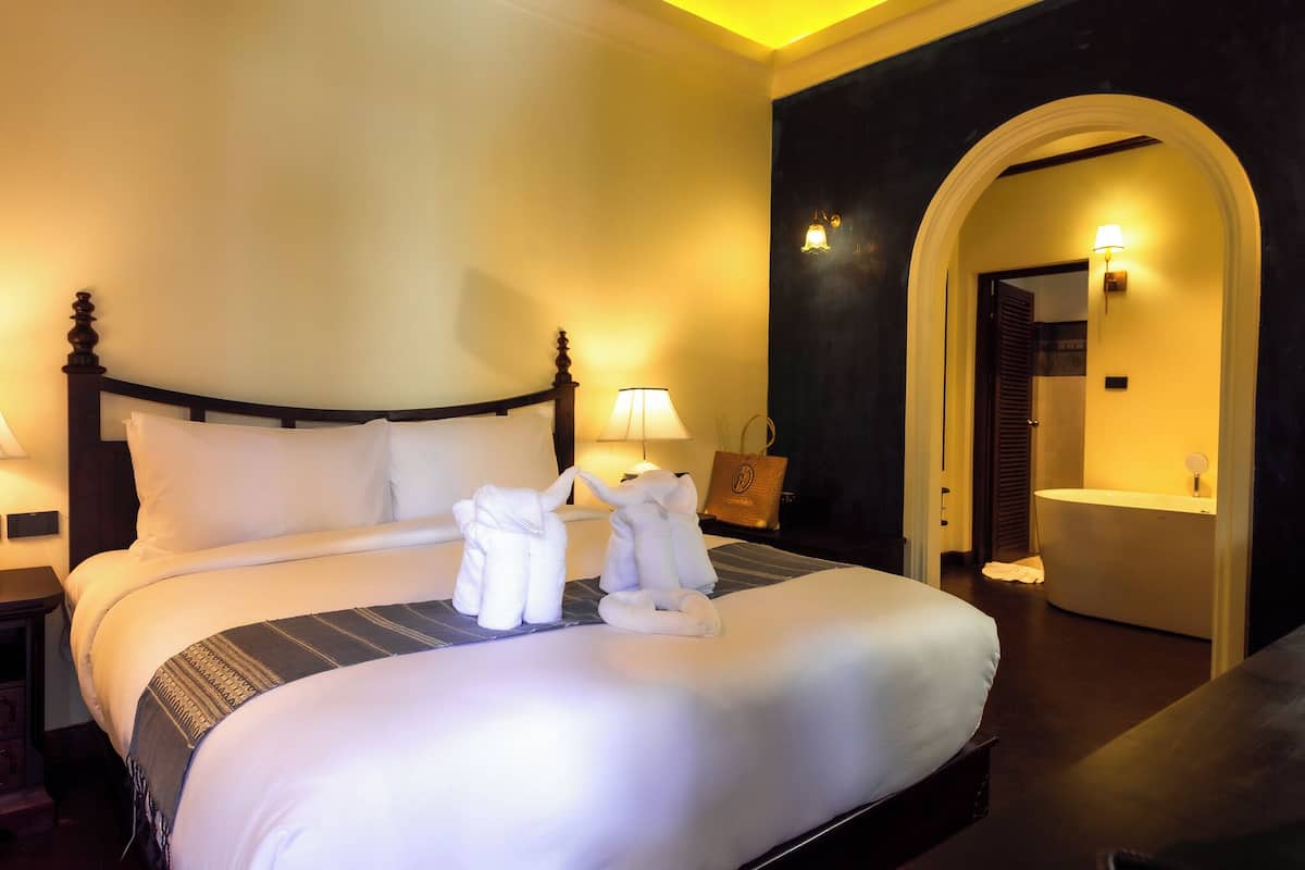 A hotel room with a large bed, a nightstand, a lamp, and a bathroom visible through an archway.
