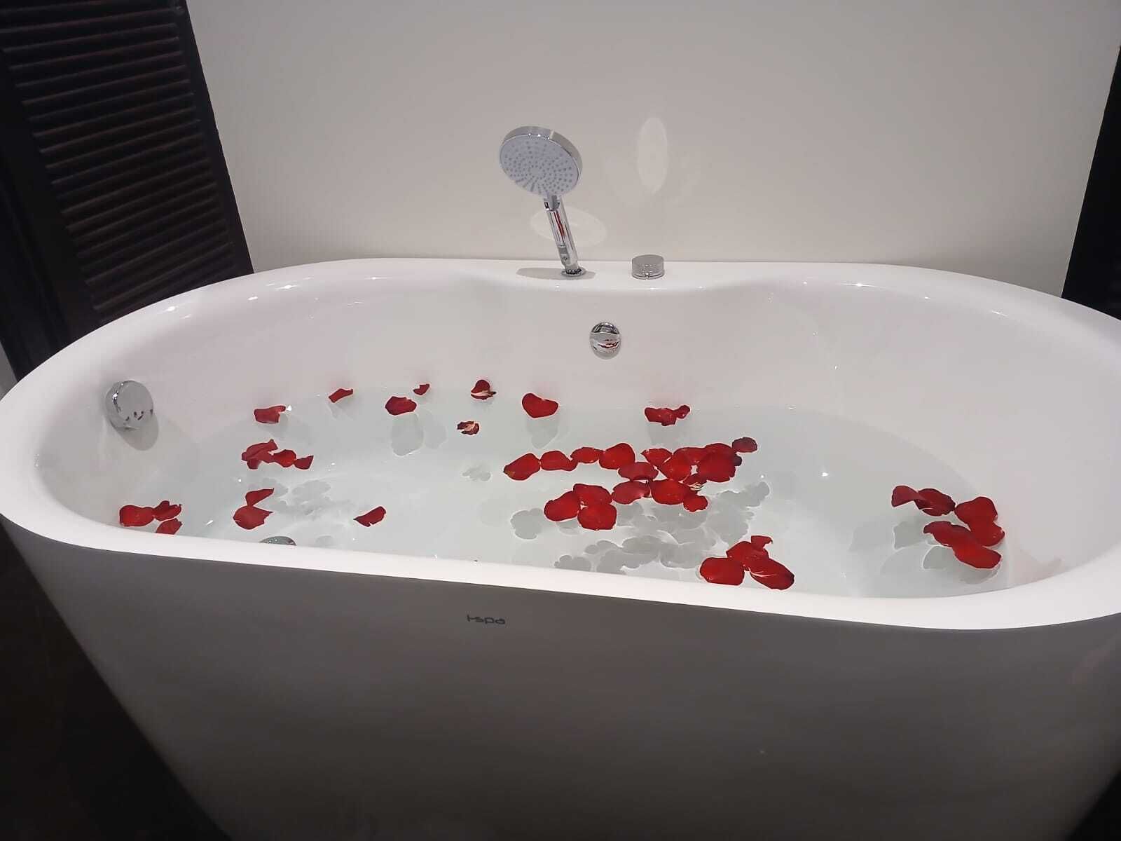 Eine weiße Badewanne mit Rosenblättern im Wasser, eine Handbrause und ein sichtbarer Abfluss.