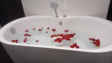 Eine weiße Badewanne mit Rosenblättern im Wasser, eine Handbrause und ein sichtbarer Abfluss.
