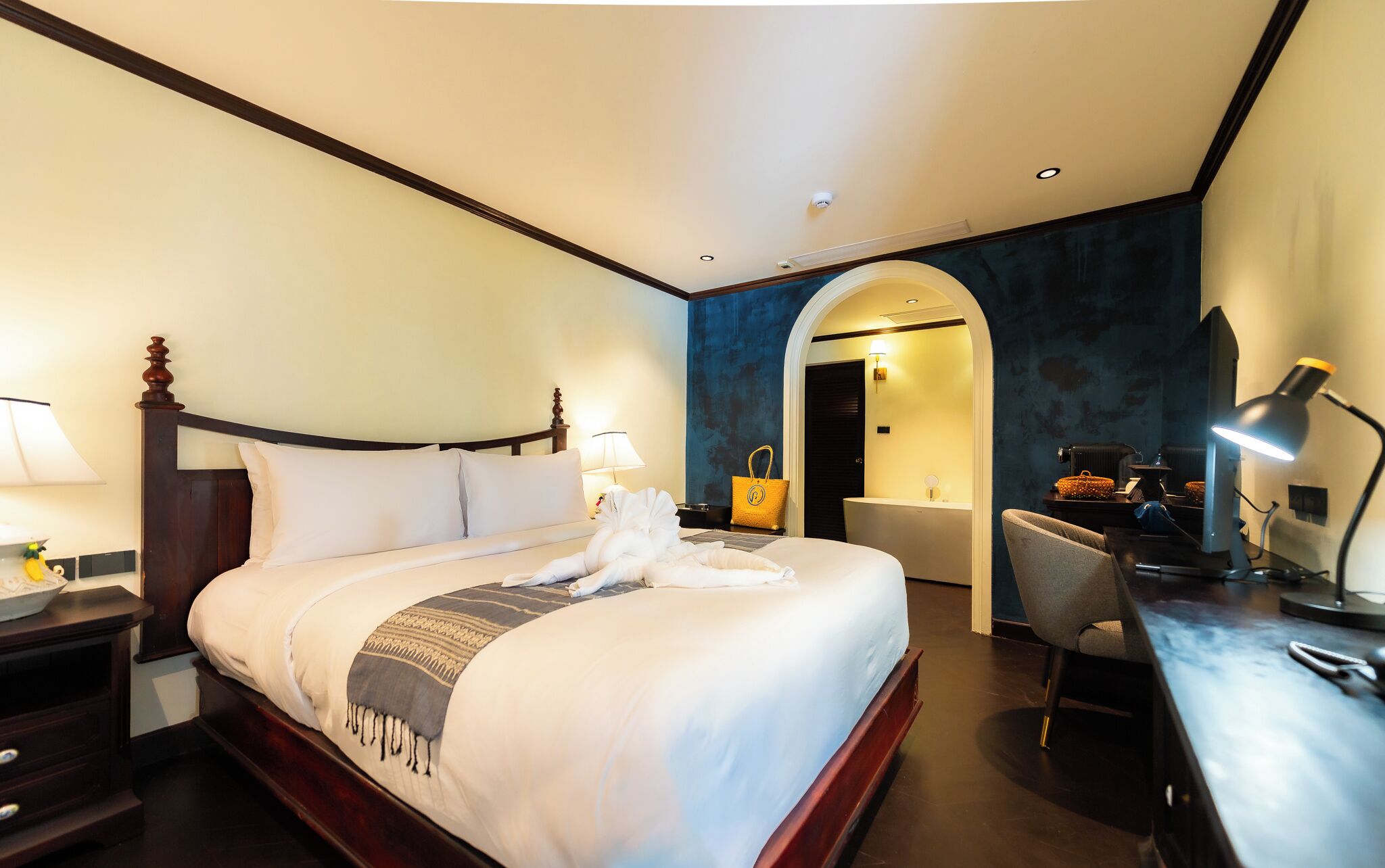 A hotel room with a large bed, a desk, a chair, a lamp, and a bathroom visible through an archway.