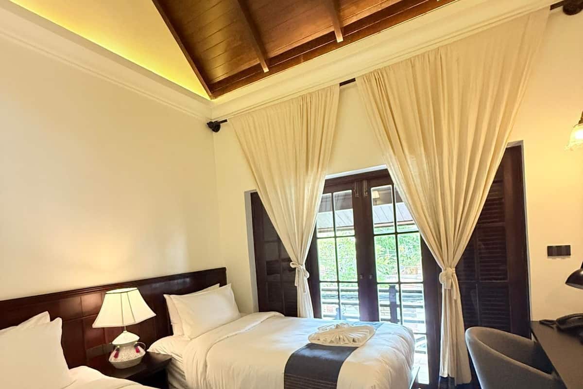 A hotel room with two beds, a desk, and a chair. There is a window with curtains and a view of greenery outside.