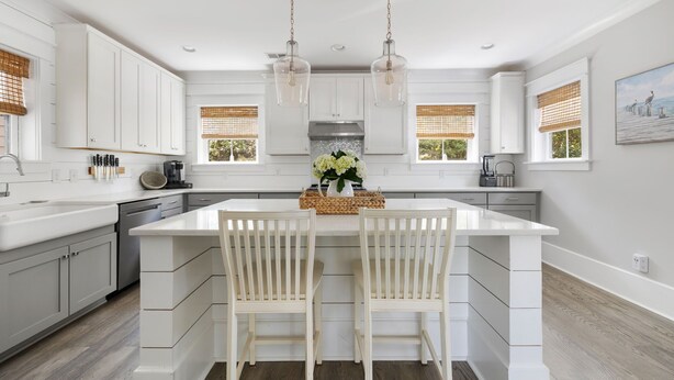 A modern kitchen with white cabinetry, a central island with two chairs, pendant lights, and a view of the outdoors through windows with blinds. - 5 Minutes to Beach|Newly Furnished Designer Home (Mount Pleasant)