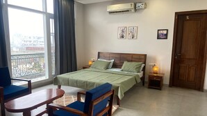 A hotel room with a large bed, two bedside tables, a wooden chair, a small table, and a view of the outside through a large window. - IT City Homestay-Secure & Comfortable Stay in Mohali (Sahibzada Ajit Singh Nagar)