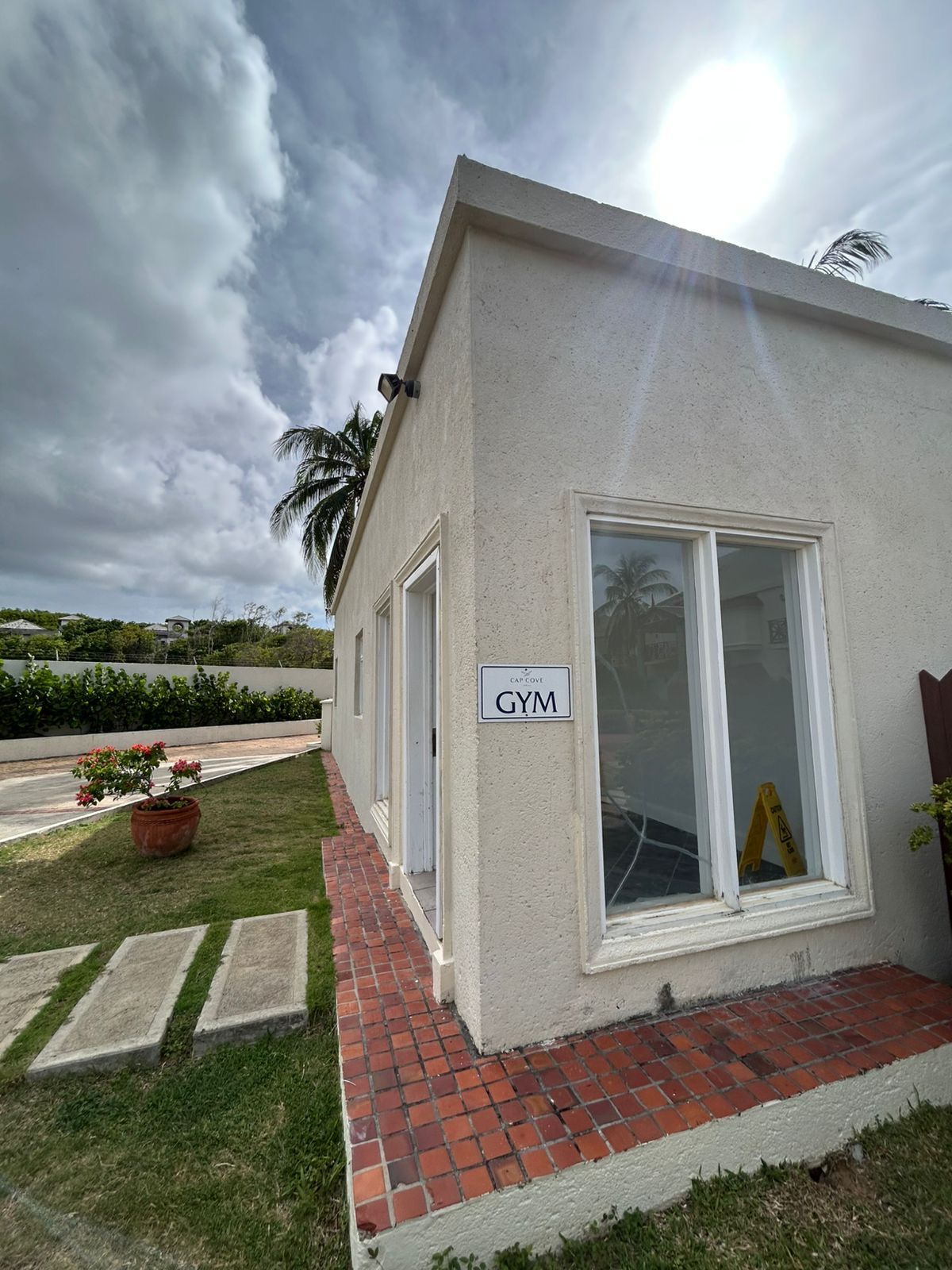 A small building with a sign reading 'GYM' on the exterior wall, a brick pathway leading to the entrance, and a palm tree in the background.