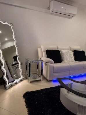 Una sala de estar moderna con un sofá blanco, almohadas blancas y negras, un espejo grande con un marco decorativo, una pequeña mesa de vidrio y una alfombra mullida negra.