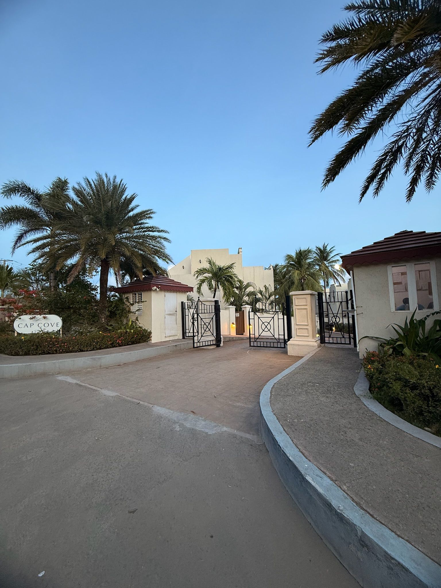 Una entrada cerrada con palmeras, un cartel que dice 'CAP COVE' y un edificio con techo rojo al fondo.