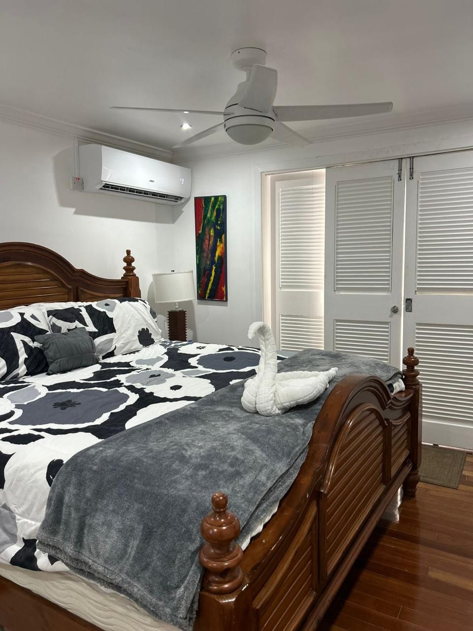 A bedroom with a bed, a ceiling fan, an air conditioning unit, a nightstand, a lamp, a painting, and a set of white doors.