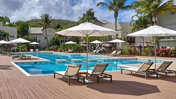 A pool area with lounge chairs and umbrellas, surrounded by palm trees and buildings. - Villa Ray: 3BR Gated Gem with Private Pool, Steps to Beach (Gros Islet)