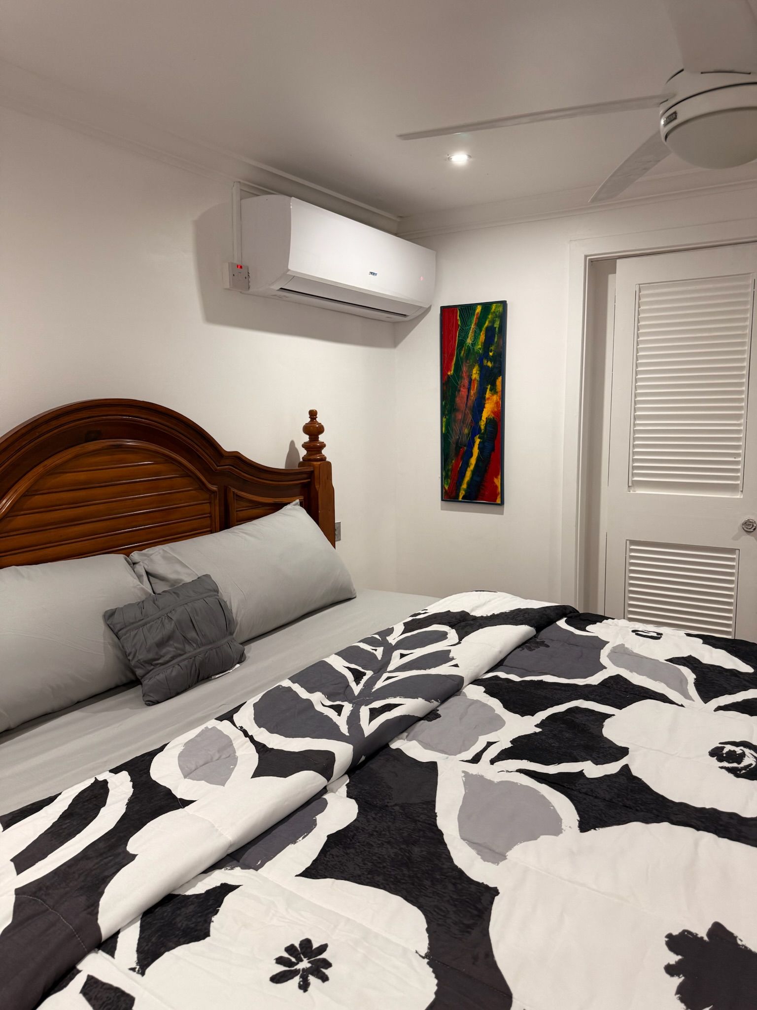 A bedroom with a wooden headboard, a bed with a patterned bedspread, an air conditioning unit, a colorful abstract painting, and a door with louvered panels.