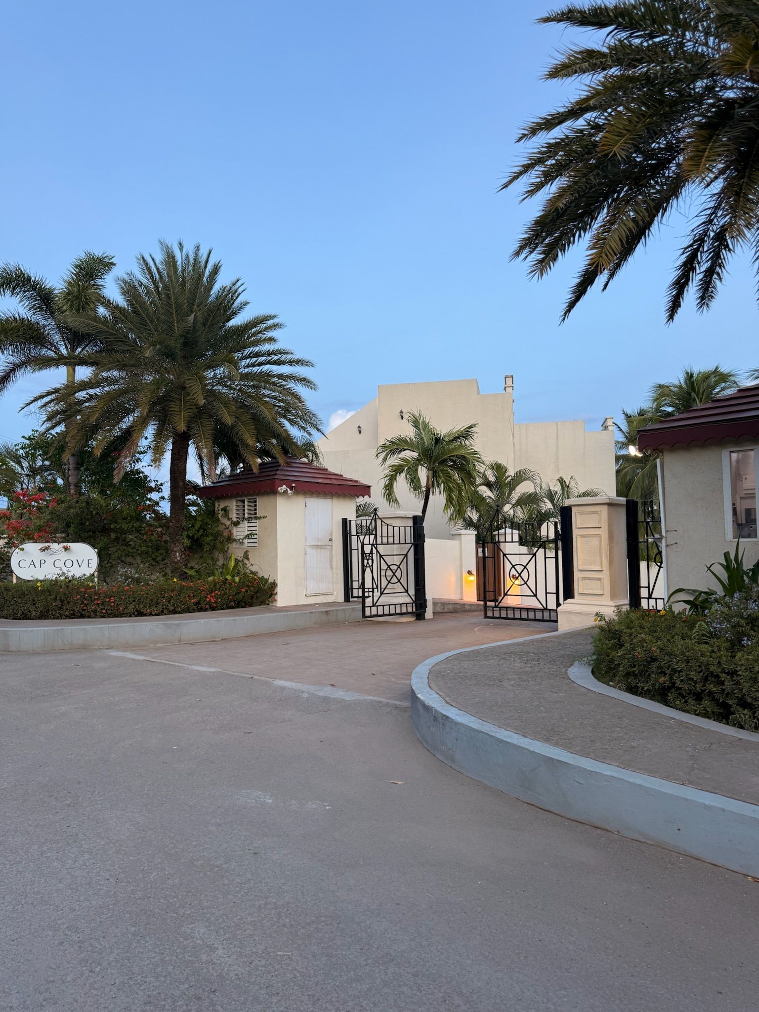 A gated entrance with palm trees, a sign reading 'CAP COVE', and a building with a flat roof.