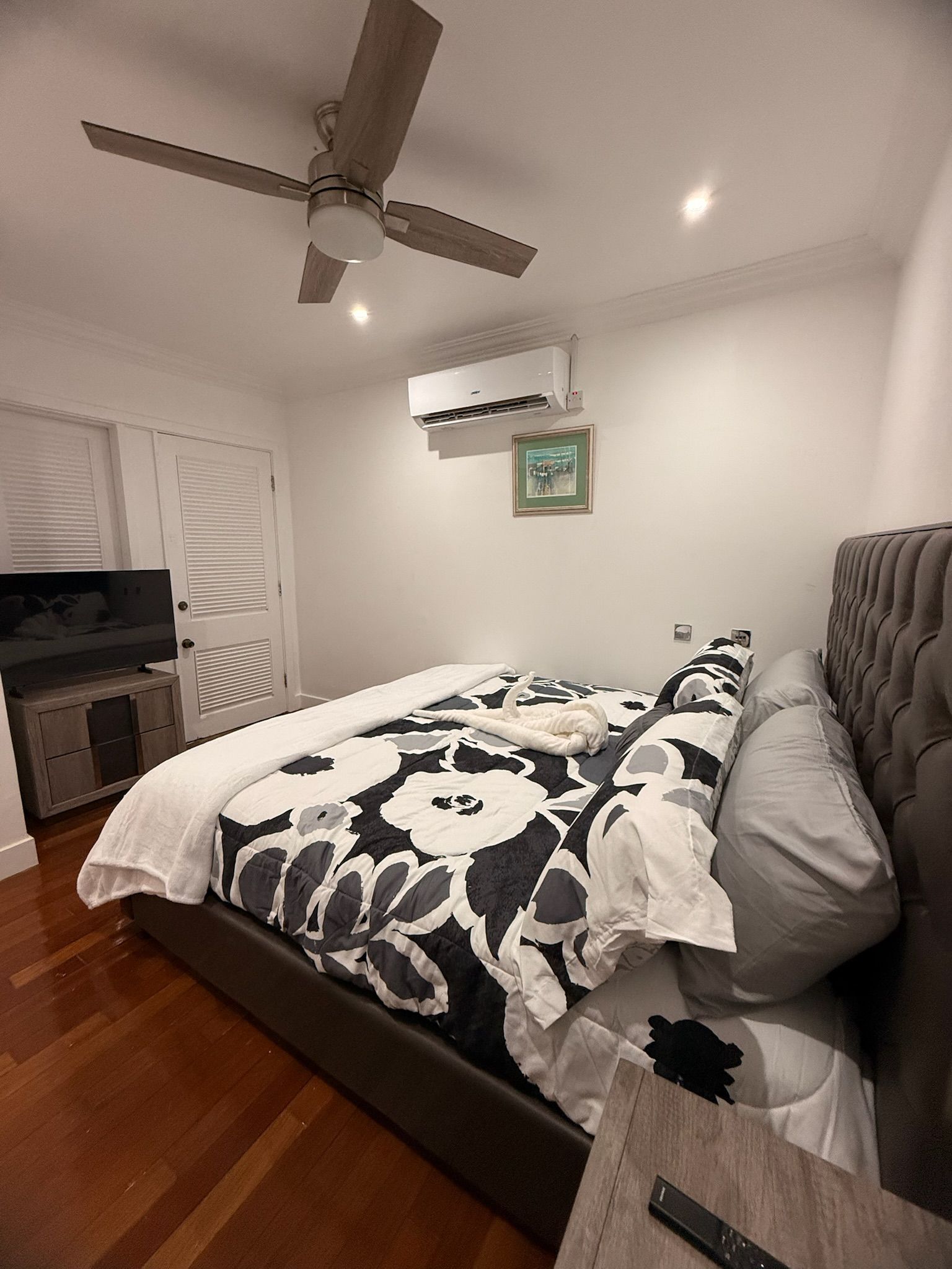 Une chambre à coucher comprenant un lit, un ventilateur de plafond, un appareil de climatisation, un téléviseur et un tableau encadré accroché au mur.