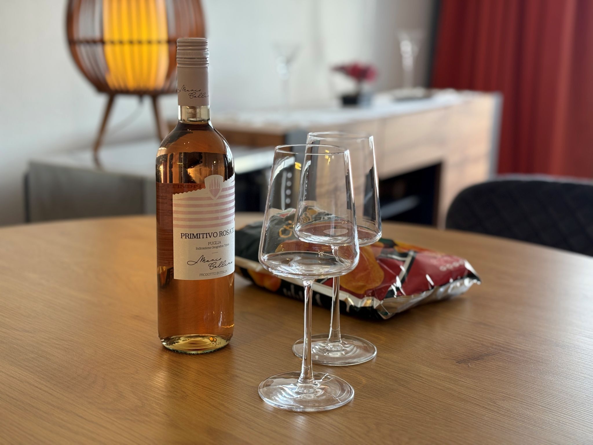 A bottle of Primitivo Rosati wine, two empty wine glasses, and a colorful bag on a wooden table.