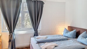 Makuuhuoneessa on kaksi sänkyä, patteri, ikkuna, jossa on verhot, ja tiiliseinä.