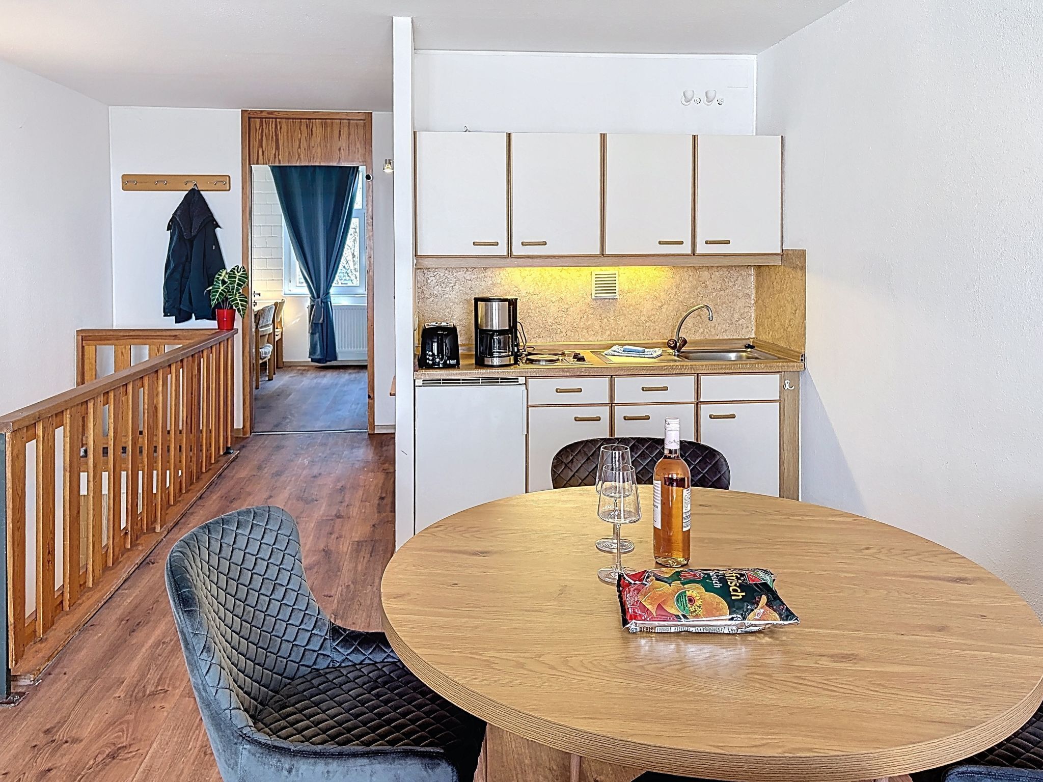 A compact kitchen with white cabinets, a coffee maker, and a sink. A dining area with a round wooden table and a cushioned chair is visible.
