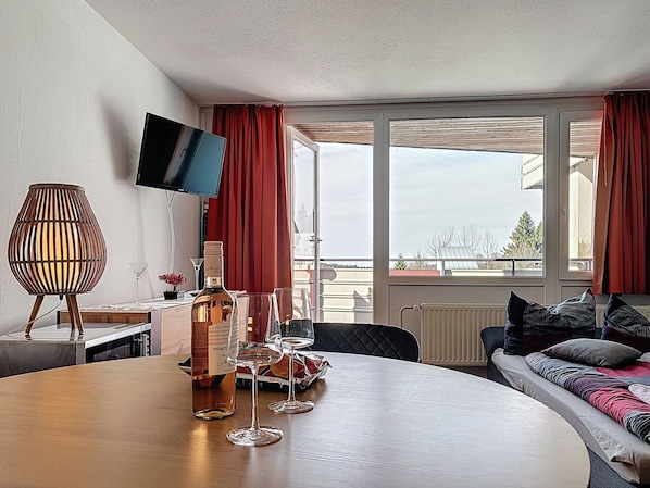 Une chambre d’hôtel avec une table de salle à manger ronde, une bouteille de vin, des verres à vin, une télévision, une fenêtre avec des rideaux et un canapé avec des coussins.