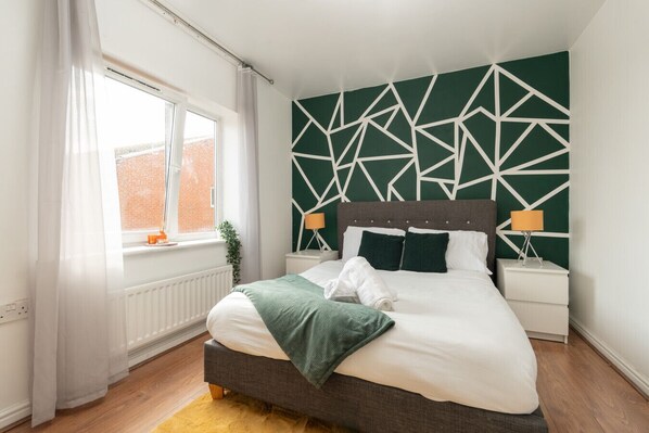 A modern bedroom with a geometric-patterned headboard, a large window with curtains, a bedside table with a lamp, and a neatly made bed.