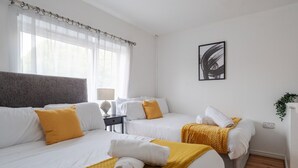 A hotel room with two beds, a bedside table with a lamp, a painting on the wall, and a potted plant.