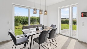 A modern dining room with a large table, grey chairs, and pendant lights. It features large windows and a view of a field.
