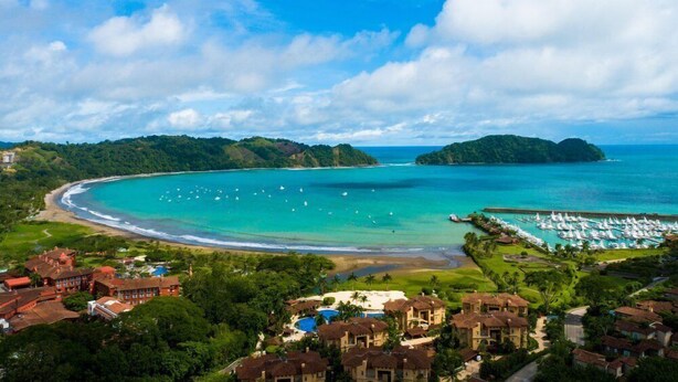 Un complejo turístico frente al mar con playa, puerto deportivo y abundante vegetación.