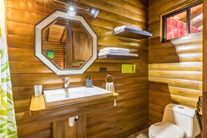 A wooden cabin bathroom with a large octagonal mirror, a sink, a toilet, and a window with towels on the wall. - Style and Comfort: Tropical Studio Cabina A/C Pool (Herradura)