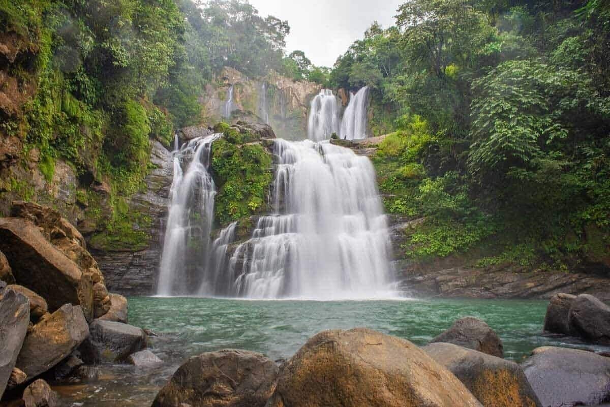 A waterfall cascading over rocks into a clear pool surrounded by lush greenery.