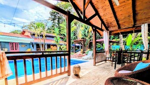 Una terraza de madera con piscina, mobiliario de mimbre y vistas a un resort tropical.