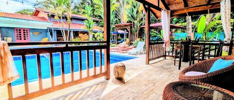 Une terrasse en bois avec une piscine, des meubles en osier et une vue sur un complexe hôtelier tropical.