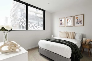 Ein modernes Schlafzimmer mit großem Fenster, einem Bett mit weißer Bettwäsche, einer dekorativen Skulptur auf einem weißen Tisch und drei gerahmten Kunstwerken an der Wand.