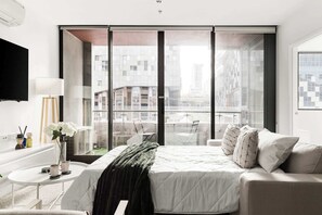 A modern hotel room with a large window, a bed, a small table, and a TV mounted on the wall.