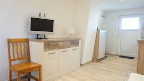 A small, clean room with a wooden chair, a TV on a wooden cabinet, and a door leading to a kitchen.