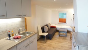 A compact kitchen with a sink, countertop, and cabinets. There is a sofa with cushions and a small table in the living area. A window with blinds is visible in the background.