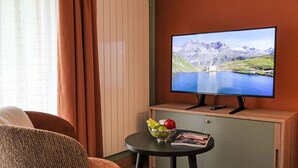 A room with a television displaying a mountain landscape, a wooden cabinet, a small table with a bowl of fruit, and a brown chair. - Beautiful apartment for 2 guests with WIFI, TV and balcony (Zermatt)