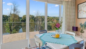 A dining area with a table set for two, a vase with flowers, and a view of a lake and trees through large windows. - Nice apartment for 4 guests with WIFI, TV, terrace and panoramic view (Vlašim)
