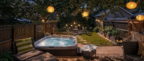 A hot tub in a backyard with a wooden deck, surrounded by outdoor furniture and string lights.