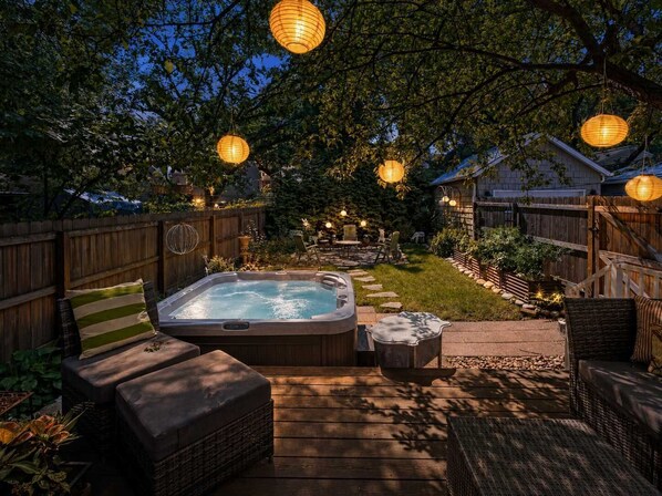 A hot tub in a backyard with a wooden deck, surrounded by outdoor furniture and string lights.