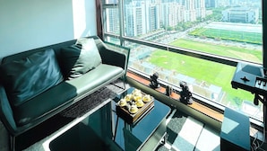 Un salón moderno con un sofá negro, una mesa de centro de cristal y vistas a un paisaje urbano a través de grandes ventanales con persianas.