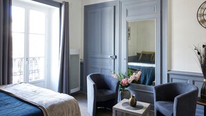 Una camera d'albergo con un letto, due poltrone, un tavolino con un vaso, uno specchio e vista su un'altra stanza.
