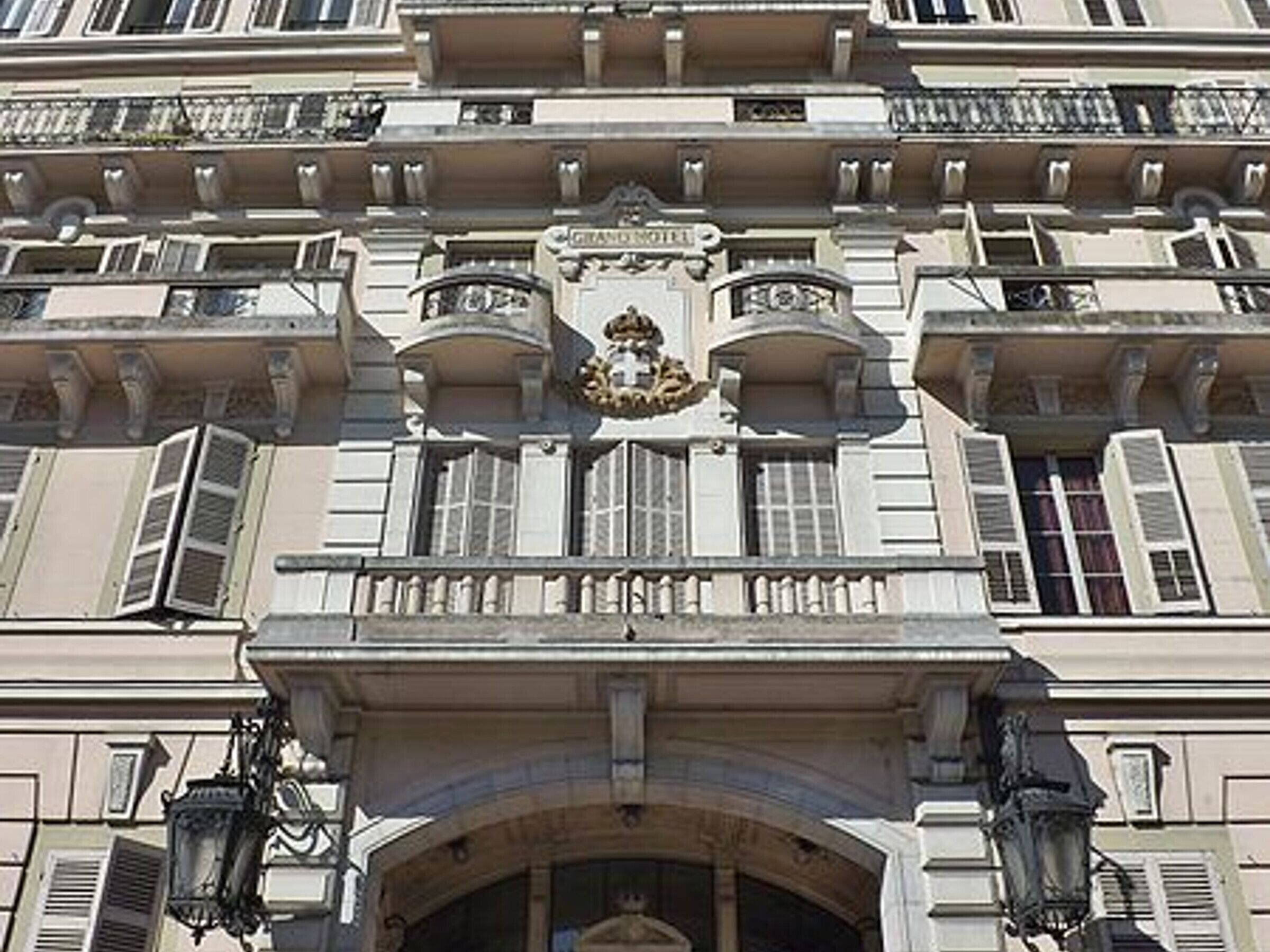 Um edifício clássico com varandas ornamentadas, entrada central com arco decorativo e brasão sobre a entrada.