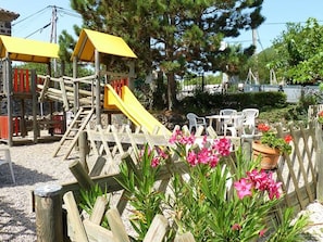 A playground with slides and climbing structures, surrounded by a wooden fence and potted plants.