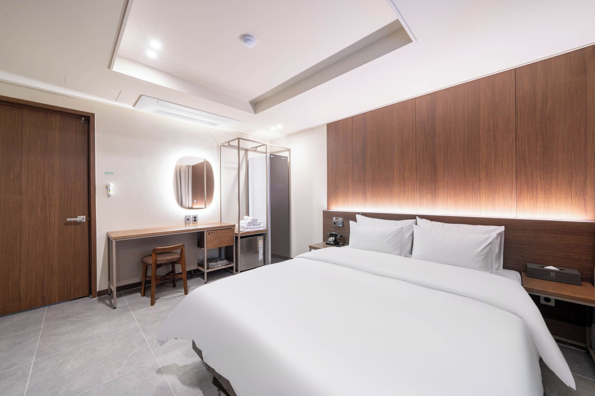 A modern hotel room with a large bed, a desk with a chair, a mirror, and a wooden accent wall.