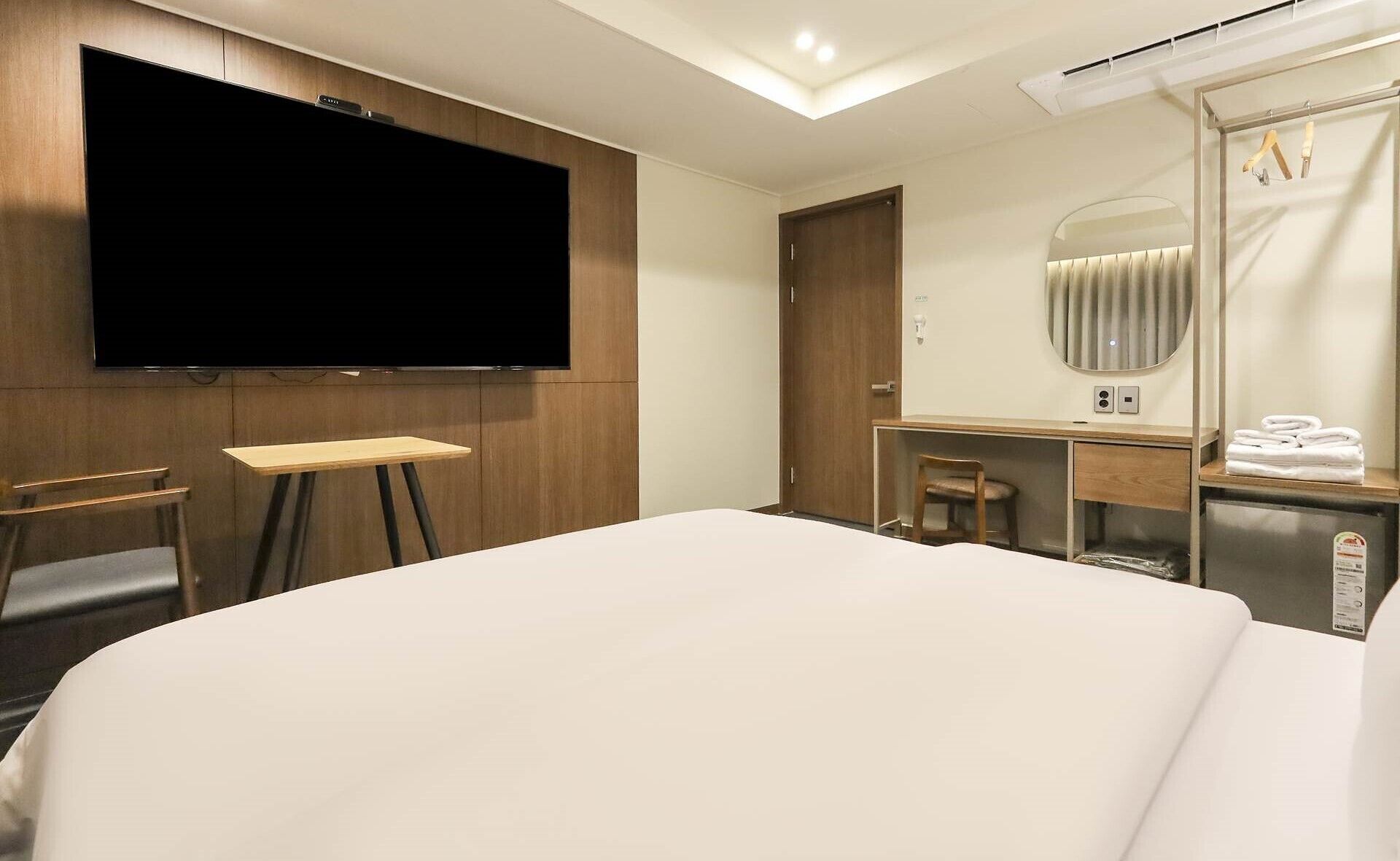 Habitación de hotel con una cama grande, televisor de pantalla plana montado en la pared, escritorio con una silla, un espejo y un estante pequeño con toallas.