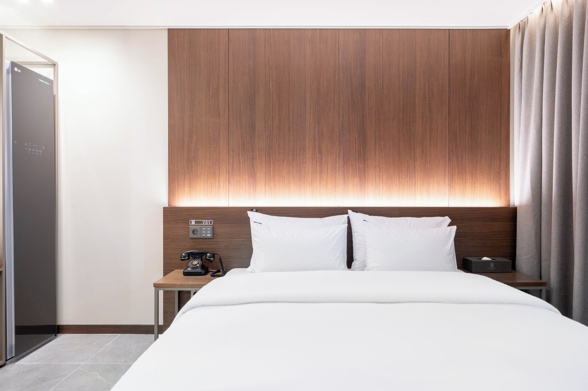 Una habitación de hotel moderna con un cabecero de madera, una cama con ropa de cama blanca, una mesita de noche con un teléfono y una caja de pañuelos, y una puerta a la izquierda.