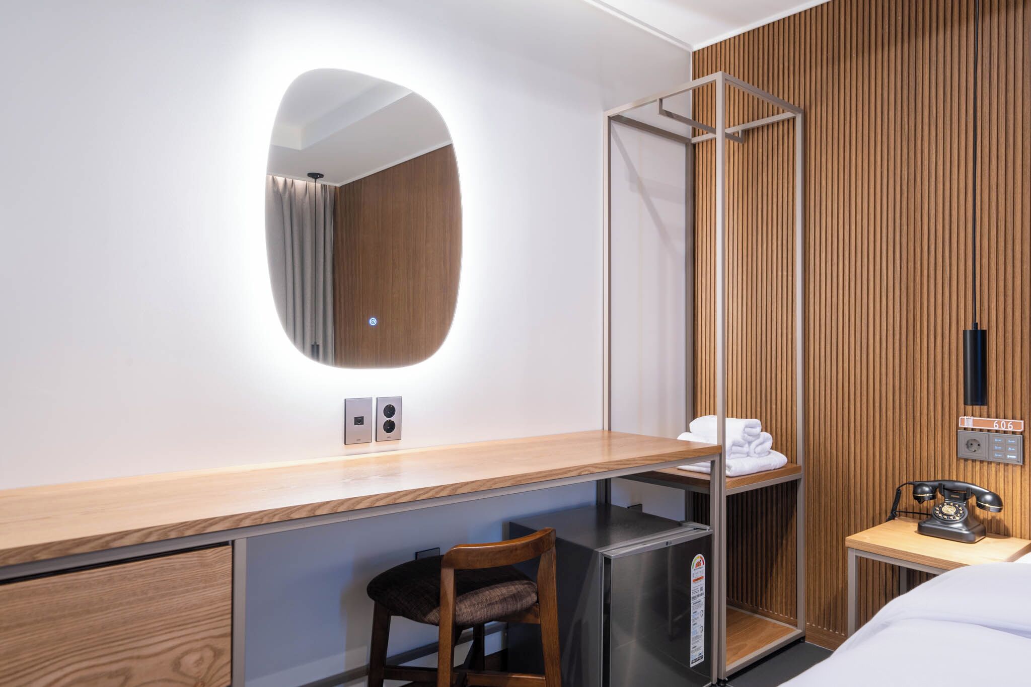 A hotel room with a wooden desk, a chair, a bed, a small shelf with towels, a mirror, a telephone, and a wall-mounted control panel.