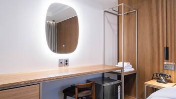 A hotel room with a wooden desk, a chair, a bed, a small shelf with towels, a mirror, a telephone, and a wall-mounted control panel.
