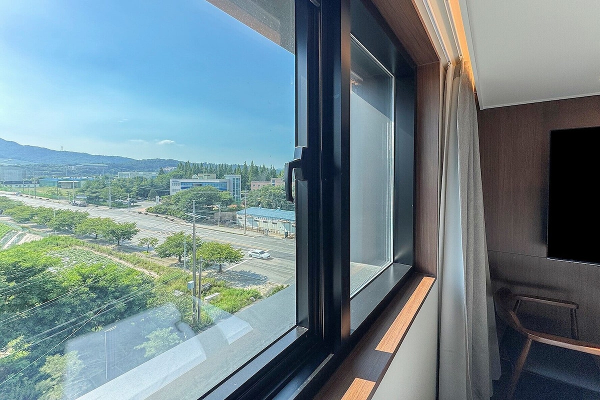 A modern room with a large window offering a city view, a flat-screen TV mounted on the wall, and a wooden bench by the window.