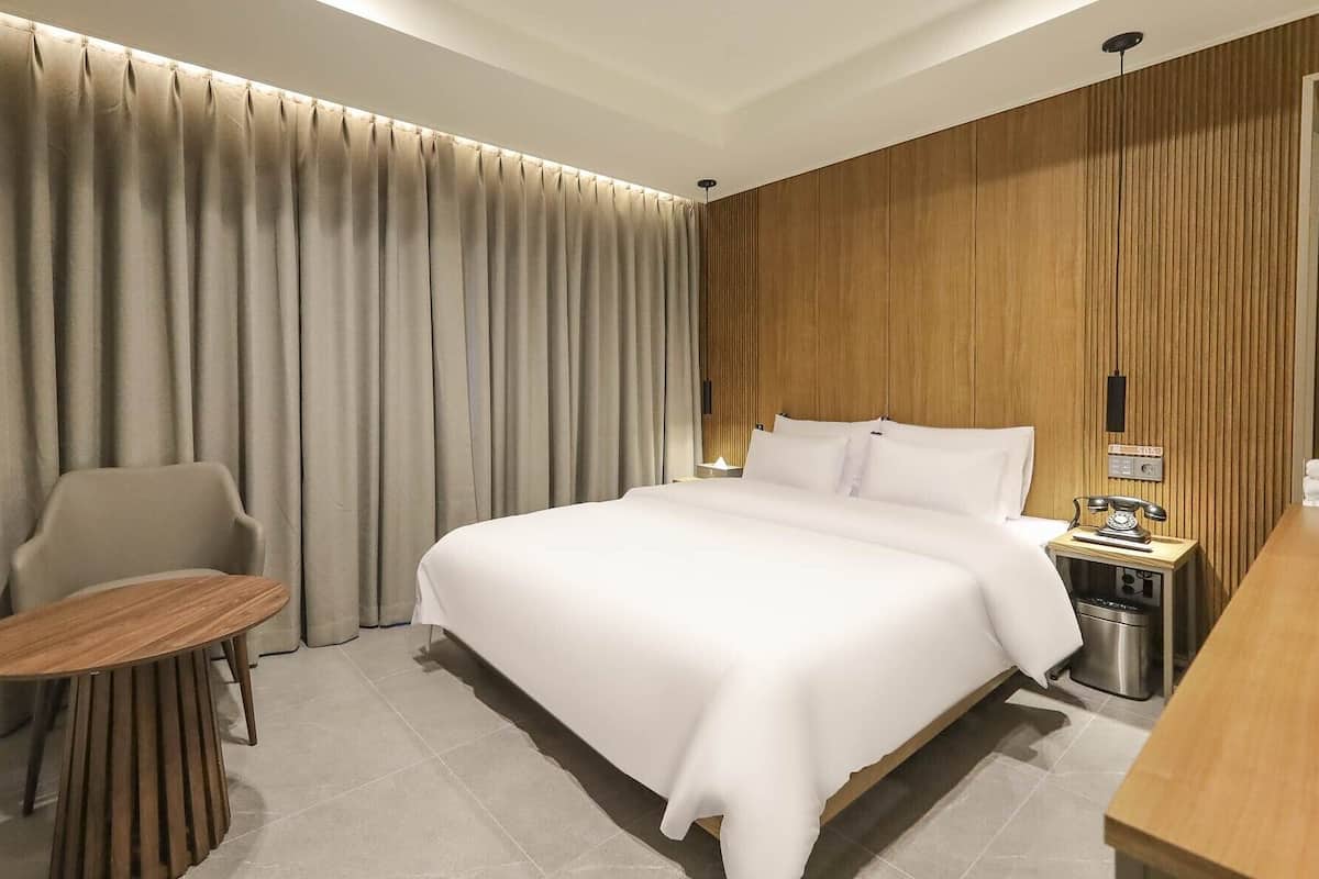 A hotel room with a large bed, a chair, a small table, and a bedside table with a lamp and a telephone.
