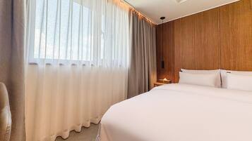 A hotel room with a large bed, a wooden headboard, a nightstand, and sheer curtains.